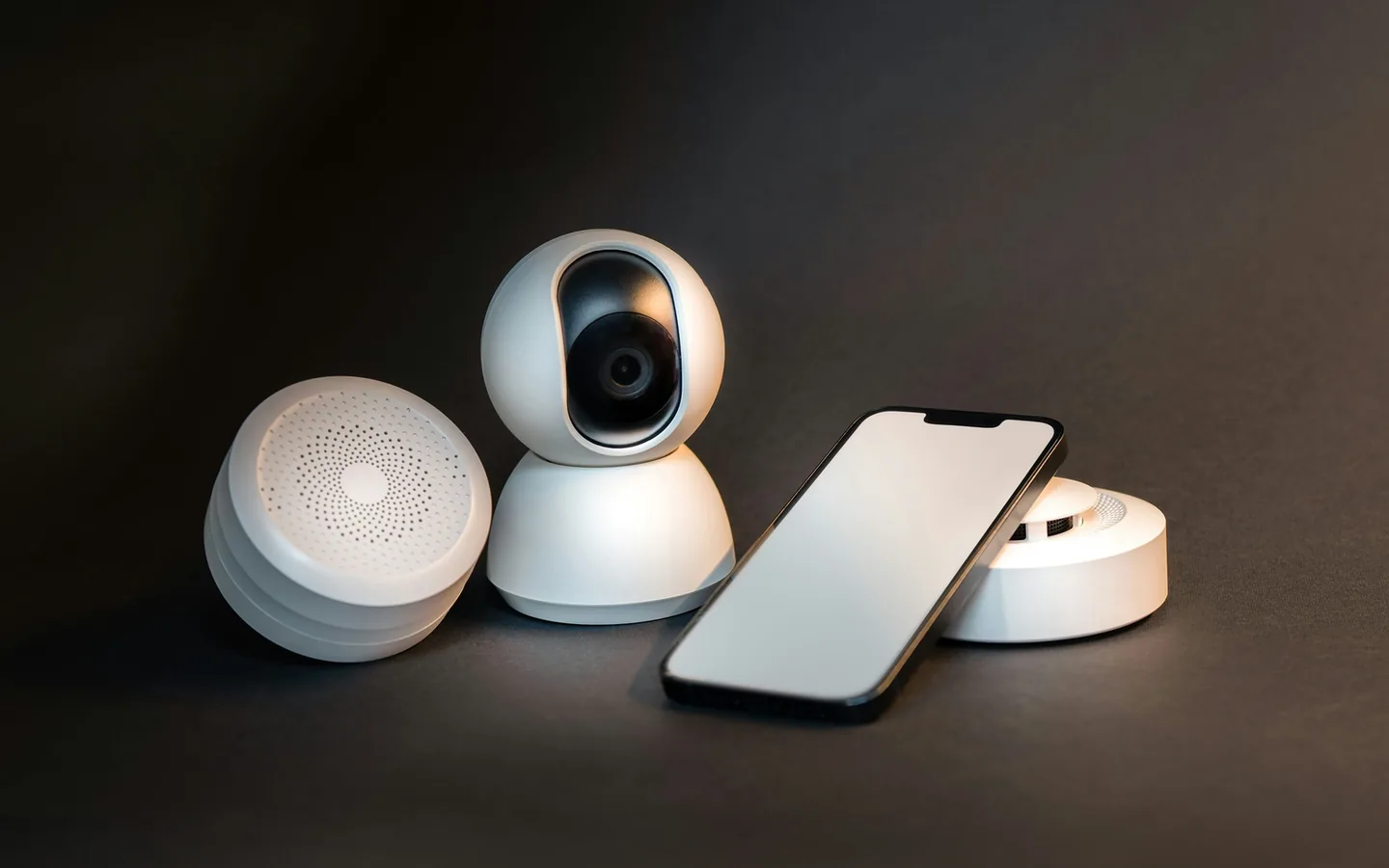 Todo aparelho conectado é também um ponto de coleta de dados. Câmera conectada de cor branca ao lado de sensores redondos e de um celular com a tela apagada, sobre fundo escuro